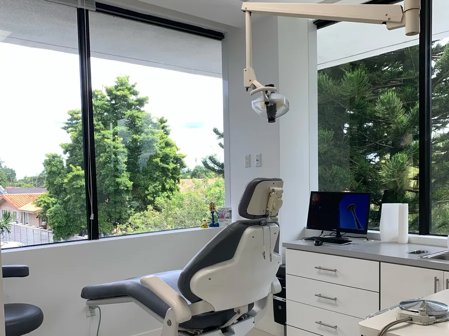 Un consultorio moderno y luminoso con un sill&oacute;n dental, un monitor de computadora y una ventana grande con vista a &aacute;rboles verdes