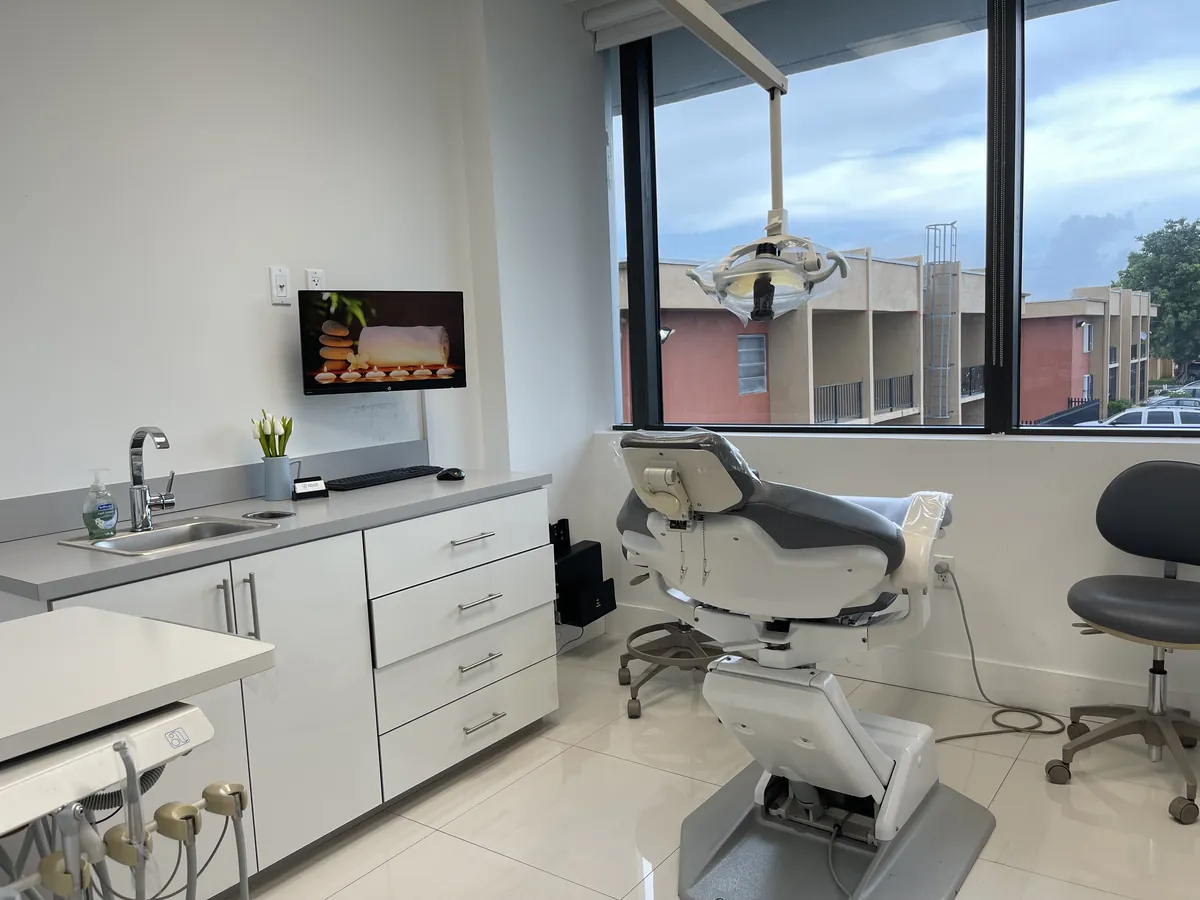 Un consultorio luminoso y moderno con sill&oacute;n dental, una ventana grande, gabinetes blancos con lavamanos y un monitor en la pared