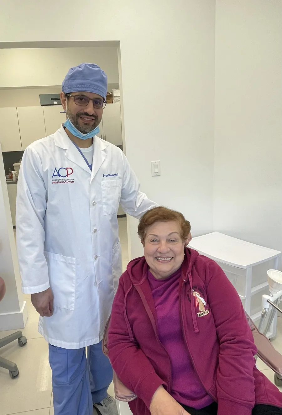 El Dr. Roy Rosado, con bata blanca y el logo del Colegio Americano de Prostodoncistas, sonriendo junto a una paciente sonriente sentada en la cl&iacute;nica