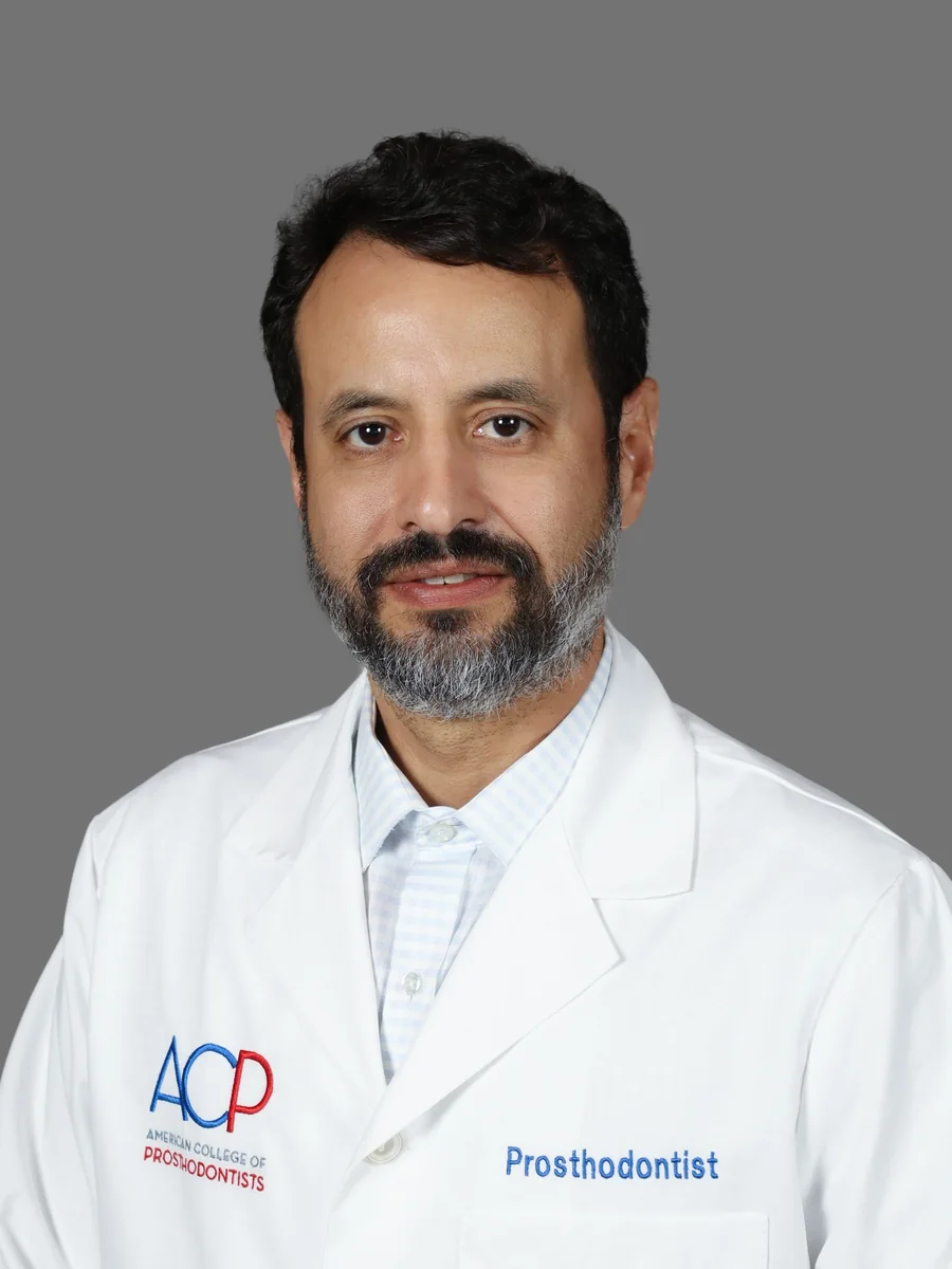 Retrato de estudio del Dr. Roy Rosado, prostodoncista, con bata blanca con el logo del Colegio Americano de Prostodoncistas y la palabra Prosthodontist bordados