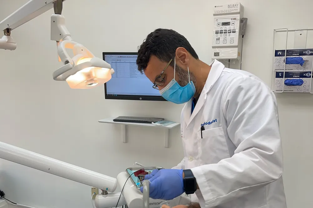 El Dr. Roy Rosado, con mascarilla, guantes y una bata blanca bordada con la palabra Prosthodontist, atendiendo a una paciente en un consultorio luminoso y ordenado