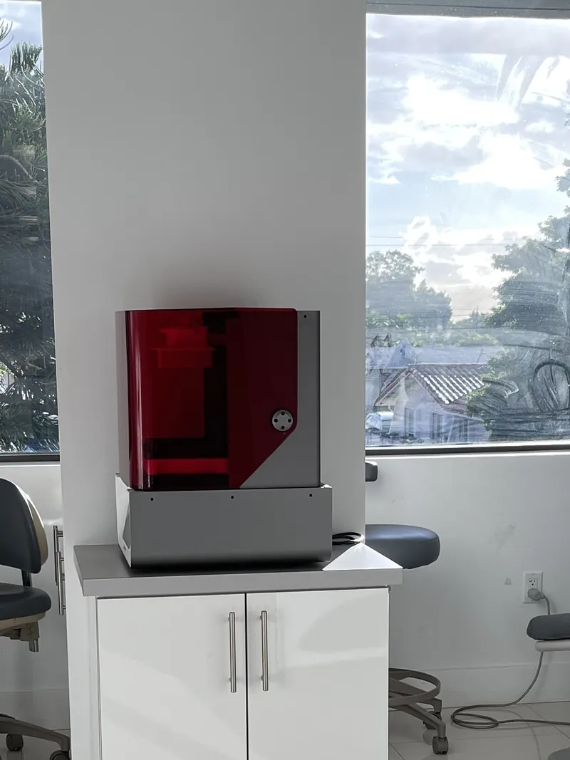 Una impresora 3D con cubierta roja transl&uacute;cida sobre un gabinete blanco en un consultorio luminoso con una ventana grande